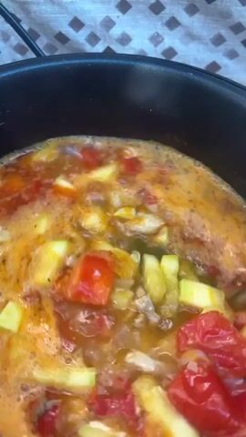 A close-up shot shows a black pot filled with a bubbling orange soup. The soup contains various chopped vegetables, including tomatoes, zucchini, potatoes, and carrots, as well as shredded chicken. A pair of green tongs is stirring the contents of the pot at the beginning of the video, and a gray spatula is used to stir the soup later in the clip. A sprig of parsley is added to the pot around the 00:01 mark. The background of the shot shows a patterned surface, possibly a tablecloth, and the edge of a black appliance.