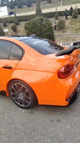 The video shows a modified orange BMW parked on an asphalt surface, possibly at a car show or gathering. The car has tinted windows, black door handles, and custom rims. There are additional black accents along the lower body. A spoiler is visible. People can be seen walking and standing on a grassy hill in the background. A banner or sign is also present. The overall environment appears to be outdoors, likely a public space during the daytime.