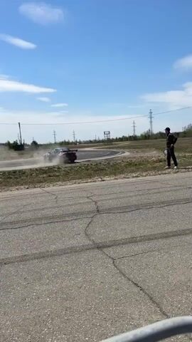 The video shows a car drifting around a corner on a racetrack. The car is dark-colored with racing stripes and a spoiler. It kicks up a lot of dust as it drifts.  A person stands nearby with a notebook, seemingly observing the action. The sky is blue with some clouds. There are utility poles and a small structure in the background, as well as some trees in the distance. Another car passes by after the drifting car is on its way out of frame. The racetrack surface is cracked and aged.