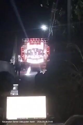 The video shows a nighttime outdoor scene, likely a road or open area, illuminated by bright lights. A large truck with a prominent sound system is the central focus. The sound system is adorned with numerous speakers and features a large, illuminated display, possibly a screen, showing a bright, cartoonish face with glowing eyes. Red lights are visible on the top of the sound system. Several people are visible, mostly silhouetted against the lights, gathered in front of and around the truck. In the foreground, a laptop is positioned, with its screen displaying what appears to be audio or video editing software. Vegetation, including tall grass or reeds, is present on the right side of the frame. A streetlamp or bright light source is visible overhead. The bottom of the frame includes a timestamp and location information: Kecamatan Semen, Kabupaten Kediri, 2023.08.28 21:44. The overall atmosphere appears to be that of an outdoor event or gathering with loud music.