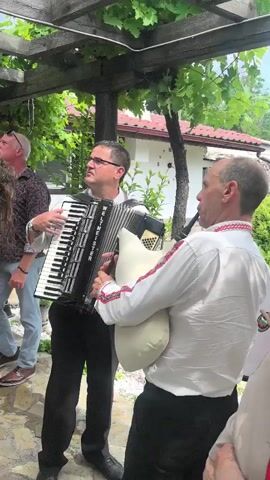 An outdoor scene features a man playing a black accordion, wearing a white shirt and dark pants, alongside a man playing bagpipes in a white embroidered shirt. A woman in a black top is partially visible to the left. The camera pans to show a bald man in a light blue shirt and glasses looking up towards a stone wall adorned with red geraniums. Above the wall, under a wooden pergola with grapevines, several individuals in white and beige clothing are visible on a raised platform, some standing, some kneeling, with one holding a white sack or fabric.