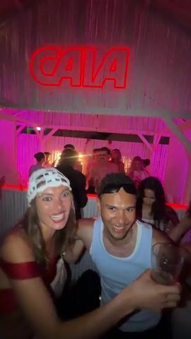 A woman with long brown hair wearing a white crocheted headpiece and a red off-the-shoulder top stands with a man in a white tank top and sunglasses. They are smiling and posing for the camera in a lively, dimly lit indoor or covered outdoor space with pink and purple lighting. A red neon sign that reads "CALA" is visible above them. Numerous other people are dancing and socializing in the background, which features white wooden paneling and a bar area. The woman has one hand on the man's chest while he has a hand near his head, possibly adjusting his sunglasses. Music plays in the background.