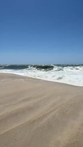 A vertical, wide shot shows a sandy beach on a clear, sunny day. The ocean stretches to the horizon under a bright blue sky. Waves crash onto the shore, creating white foam that recedes across the sand. The water is a deep blue-green, and the waves appear to be quite large, with some cresting and breaking dramatically. The sand in the foreground is light brown and appears dry, with subtle patterns formed by the wind. There are no people or other objects visible in the scene.