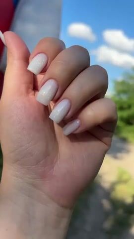 This close-up media file features a person's hand with neatly manicured nails. The nails are painted a shimmery white with iridescent glitter, cut in a square shape with rounded edges. The hand is held up against a backdrop of a bright blue sky with fluffy white clouds, suggesting a clear day. Lush green foliage and trees are visible in the background, hinting at an outdoor setting. A blurred red and white object, possibly a flag or part of a structure, is partially visible on the far left. The sunlight suggests it is daytime, likely late morning or afternoon. The overall impression is one of simple elegance and enjoyment of the outdoors, possibly during a warm season in Zubov, Russia. There is no visible text in the media file.