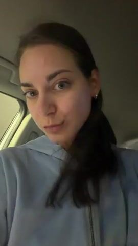 A woman is in the backseat of a car. The camera is pointing at her. She is wearing a light blue, zip-up hoodie and has her brown hair in a ponytail. She looks directly at the camera with a neutral expression for several seconds, then smiles at the camera. The interior of the car is visible. The front passenger seat is visible, as well as the back seats. The environment outside the car is dark, with glimpses of streetlights.