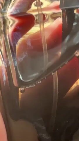 The video features a close-up shot of a clear container, likely glass or plastic, holding a dark liquid, possibly a beverage. A clear plastic straw or tube is submerged in the liquid. Reflections of a reddish-brown object and other bright, textured surfaces are visible on the curved surface of the container, shifting with slight movements. Small bubbles can be observed rising through the dark liquid, particularly near the base of the straw at the 0:01 mark. The background is out of focus, emphasizing the container and its contents. No people or text are explicitly visible.