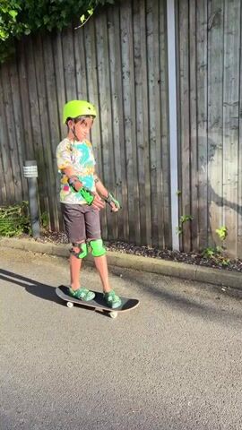 A young boy wearing a neon green helmet, elbow pads, wrist guards, and knee pads stands on a skateboard. He is wearing a t-shirt with a colorful graphic and black shorts. The skateboard is on an asphalt surface next to a wooden fence. The sun is shining, casting shadows. The boy appears to be preparing to skate.