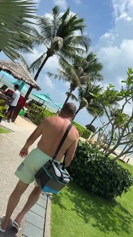A person, seen from behind, walks along a paved path on a sunny day. They are wearing light-colored swim trunks and have a dark and blue bag slung over their shoulder. The path is bordered by green grass and lush foliage. In the background, palm trees with green fronds sway under a partly cloudy blue sky. To the left, a small thatched-roof hut with a counter is visible, with a person standing behind it. Beyond the hut, beach chairs and umbrellas are set up on a sandy beach leading to the ocean. The overall scene depicts a tropical resort setting.