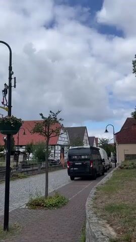 The video opens on a street scene with a black van parked on the left side of the road. The road is paved with cobblestones, and there are traditional German half-timbered houses lining the street. A brick sidewalk runs along the side of the road, bordered by a low stone wall and a grassy area. A small tree grows on the left side of the sidewalk. The sky is cloudy.

The camera then pans to the right, revealing a sculpture of six women sitting on a red sofa in a grassy park area. The women are depicted in various poses and appear to be wearing matching pinkish-red dresses and shoes. They are all smiling. Behind the sculpture, there are large trees, and in the distance, there are more houses. A white signpost with a directional arrow is visible in the upper right corner of the frame. The lighting suggests it is daytime.