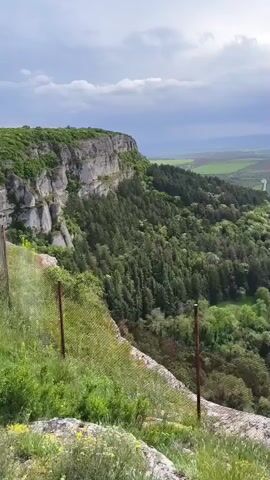 The video shows a panoramic view of a landscape from a high vantage point. The foreground consists of a rocky outcrop with green vegetation and a chain-link fence. Beyond this is a dense forest that slopes downward. In the distance, there is a view of flat fields, some of which are yellow, possibly due to crops. A small town or settlement is visible nestled amidst the greenery. The sky is overcast with a mix of dark and light clouds, suggesting varied weather conditions. There are no people visible in the video. The camera pans slightly to the right, providing a broader view of the scene. The focus is on the natural landscape and the town in the distance.