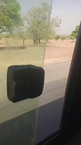 The video is shot from inside a moving vehicle. The view through the side window reveals a rural landscape with scattered trees and a brown field, suggesting an arid environment. A paved road runs alongside the field. A small, dark, rectangular object is attached to the window, partially obstructing the view. As the vehicle moves, a few people can be seen standing on the side of the road near a car, but they are somewhat blurry. The overall impression is that the video was taken during a trip through a dry, rural area.