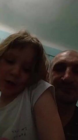 A man is holding a small child in front of a camera. The girl is wearing a white t-shirt and has shoulder-length blond hair. The man is bald. They are both looking at the camera. The girl raises both hands and extends her middle fingers. A window with a sheer curtain can be seen in the background. The wall and ceiling are painted in a light blue or turquoise color.