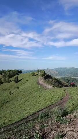 The video pans across a rolling green hillside with a dirt path winding along its crest. Scattered trees dot the landscape. The sky is a vibrant blue with scattered white clouds. In the distance, a valley opens up with more rolling hills and a body of water. The scene appears to be a natural, outdoor environment, possibly a park or scenic overlook, with no people or specific objects of interest visible. The overall impression is one of natural beauty and open space.