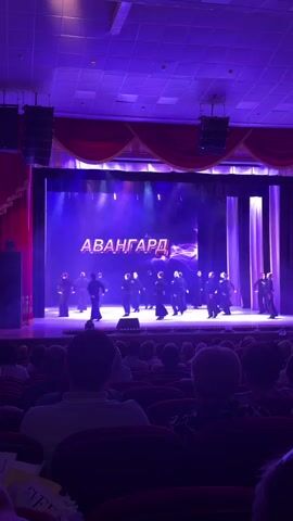 The video shows a wide shot of a stage performance inside an auditorium. On stage, approximately a dozen male dancers in dark costumes are performing a synchronized routine. Behind them, a large screen displays the word "АВАНГАРД" (AVANGARD) in red and white letters, with a purple swirling graphic in the background. The stage is illuminated with purple and white stage lights. Red curtains frame the sides of the stage. In the foreground, the backs of several audience members are visible, seated in rows of red chairs, watching the performance. Two large black speakers are mounted on either side of the stage.