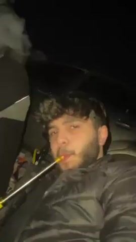 A man with a beard and curly hair, wearing a black puffer jacket, is shown smoking a hookah. He is in a dark environment, possibly a car, with another person partially visible in the background. The man takes a drag from the hookah, exhaling a large cloud of white smoke. The camera focuses on the smoke as it billows out and obscures the view.