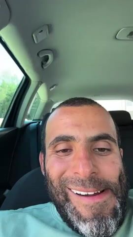 The video is a close-up, selfie-style shot of a man's face. He is sitting in the driver's seat of a car. The man has dark hair and a beard with some gray. He is smiling broadly, with his mouth open and teeth visible. His eyes are slightly closed, and he appears to be looking directly at the camera. The interior of the car is visible behind him, including the ceiling with lights and the back seat. The lighting suggests it is daytime, and the man is outdoors, as indicated by the greenery visible through the car window.
