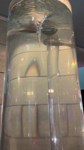 A cylindrical glass tank is filled with water. A vortex is forming in the center of the tank, with water spiraling downwards into a central tube. The tank is illuminated, creating reflections and refractions on its surface. The background appears to be an indoor environment with tiled walls and some text visible on them. The overall scene captures the dynamic nature of fluid dynamics.