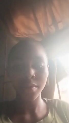 The video shows a close-up selfie of a young Black person with short hair. They are wearing a green t-shirt. The person appears to be indoors, with a window and curtains visible in the background. The lighting suggests it is daytime, with sunlight coming through the window, creating some glare. The person is animated and smiling, looking directly at the camera. They speak briefly in English, saying "Jim jim guys, welcome back back, yo yo yo." The audio is clear. The facial expressions suggest a happy and welcoming demeanor. The setting is likely a home in Afaha Ukwa, Nigeria.