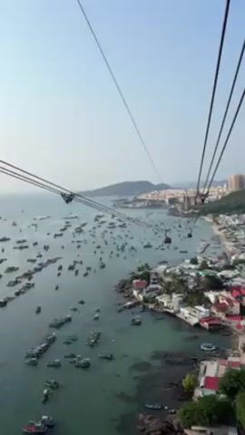 The video shows an aerial view of a coastal area with many boats in the water, taken from a cable car. The cable car's wires are visible in the foreground.  The water is a clear turquoise color.  There are buildings on the coast, including some red-roofed structures. In the background, there is a town with taller buildings on a peninsula. The sky is bright blue and clear.