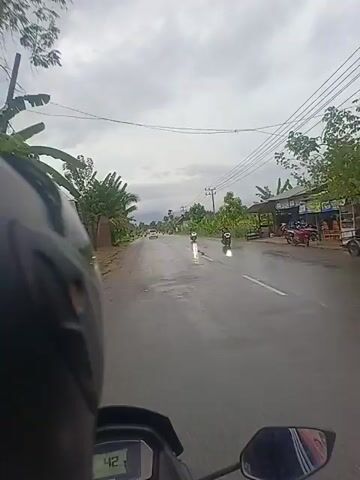 A first-person perspective video captures a motorcycle ride along a wet, two-lane asphalt road under an overcast sky. The road appears recently rained upon or is experiencing light drizzle, with puddles and reflections visible.

In the foreground, the edge of the rider's black helmet is visible on the left, and the dashboard of the motorcycle is partially seen at the bottom, displaying the number "42".

The road stretches into the distance, with a white car briefly visible far ahead. Several other motorcyclists are seen on the road; one passes on the left, displaying an Indonesian license plate "BL 3072 FAP".

The roadside is lined with lush green vegetation, including palm trees and what appear to be banana plants. Simple, single-story structures or shacks are scattered along the right side of the road. Overhead, power lines run parallel to the road, supported by wooden poles. The overall impression is a rural or semi-rural setting in Indonesia, consistent with Cotentung. The weather is gloomy, with a heavily clouded sky.