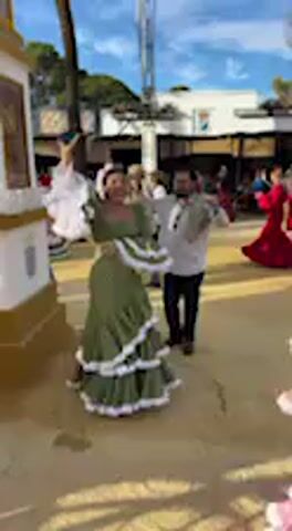 The video depicts a vibrant outdoor scene at what appears to be a traditional Spanish "Feria," likely in Jerez de la Frontera, Spain, given the cultural context. The setting features a sandy or dirt ground area, indicative of a fairground, with various people, temporary structures, and buildings in the background under a bright blue sky with some clouds, suggesting pleasant daytime weather.

The main activity is traditional Spanish dancing, likely Sevillanas. In the foreground, a woman wearing an olive-green flamenco dress with white ruffles and a white top dances with a man dressed in a light-colored shirt and dark trousers. Both are moving energetically and appear joyful. The woman raises her arms gracefully, showcasing her dress.

As the camera shifts, another woman enters the frame, captivating in a pink flamenco dress adorned with white polka dots and multiple tiers of ruffles, paired with a white fringed shawl. She also engages in the dance, turning and posing with elegance. In the background, other attendees, some dressed in similar elaborate flamenco dresses (including red, white, and black), are seen moving, watching, or participating in the dancing, contributing to the lively and festive ambiance. The scene evokes a strong sense of cultural celebration and community engagement. There is no visible text in the video.