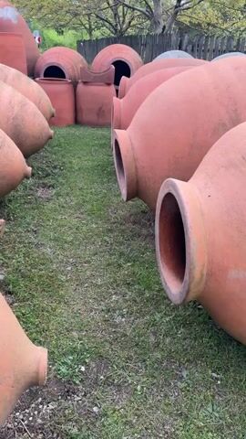 The camera captures a collection of large, terracotta-colored clay pots and spherical structures resting on a patch of green grass. The pots are arranged in rows, with openings facing the camera, revealing their hollow interiors. Behind them are several spherical structures, some with circular openings. Further in the background, a weathered wooden fence is visible, partially obscured by trees with bare branches. The scene is outdoors, with overcast lighting.