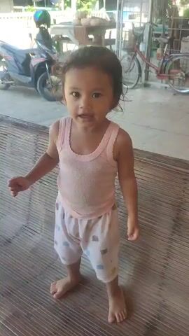 A young child, appearing to be a toddler, stands on a slatted surface, likely a bench, and smiles at the camera. The child is wearing a pink tank top and white shorts with a pattern. The child's hands are positioned to the sides of their body. In the background, a motorcycle is parked, and further behind, a bicycle is visible. The setting appears to be outdoors, possibly a yard or patio area, with some structures and vegetation in the distance. The child's demeanor is cheerful and engaging.