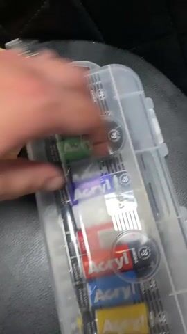 The video shows a hand opening a clear plastic container filled with tubes of acrylic paint. The tubes are arranged in rows and appear to be of various colors, with labels that include the brand name "Acryl" and "Primo." The container is placed on a dark, textured surface, possibly a car seat. The hand is seen reaching into the container and manipulating the latch to open it. The lighting is dim.