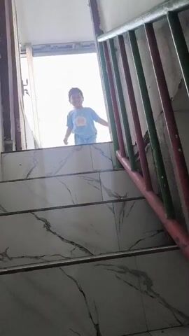A young child stands at the top of a staircase, looking down at the camera. The child is wearing a blue t-shirt and shorts. The staircase has marble-like tiles on the steps and a metal railing with vertical bars. The background is brightly lit, suggesting an open doorway or window. The child smiles and briefly raises their hands to their head before lowering them. The video is short, with the child's actions repeated slightly.