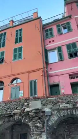 The video starts with a low-angle shot of brightly colored buildings with green shutters, built on a stone foundation. The building colors visible include yellow, red, pink, and orange. The camera then pans upwards to show the clear blue sky and the sea. The sea is calm, and a rock barrier extends into the water. There are several buoys floating in the water near the barrier. In the distance, people are standing on the rocks at the end of the barrier.  A plane's contrail is visible high in the sky.