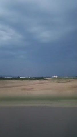 The video shows a landscape with a cloudy sky, a field of grass and vegetation, and a road in the foreground. The lighting suggests it is either dawn or dusk, or that a storm is approaching. There are no people or obvious man-made objects visible in the distance, except for what appears to be a distant structure or lights. The camera is moving, indicating that this footage was likely captured from a vehicle. The overall atmosphere is somber and somewhat dramatic due to the stormy sky.