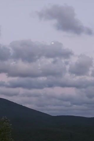 The video shows a dusky sky with scattered clouds. The moon is visible through a gap in the clouds, and it appears to be a crescent or gibbous moon. Below the sky, a dark, tree-covered mountain range stretches across the frame. The lighting suggests that it is either dusk or dawn.