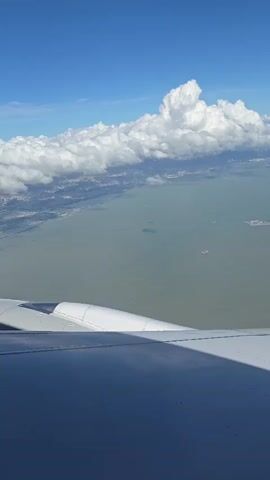 The video shows an aerial view from an airplane window looking out over a large body of water and a distant coastline. The airplane's wing, a white curved surface with a shadow cast on it, occupies the bottom portion of the frame. The water is a muted green-gray color and appears relatively calm, with a few small boats visible. In the distance, a long bridge, with a distinctive cable-stayed section, spans across the water, connecting to land on both sides. The coastline is dotted with buildings and structures, suggesting an urban or industrial area. The sky is a vibrant blue, with scattered white cumulus clouds. One large cloud formation stands out prominently above the distant landmass. The overall impression is a clear, sunny day with good visibility.
