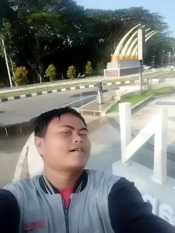 A man takes a selfie in front of a large white sculpture and a monument of a muscular figure holding a club. The man is wearing a gray and black jacket over a red shirt. He has short dark hair and is looking directly at the camera. In the background, there is a large white sculpture with the word "NGAWI" visible. Behind the sculpture, there is a monument depicting a muscular figure standing on a stone base, holding a large club. Further back, there is another monument featuring several white tusks arranged in a fan shape. The scene is outdoors, with a road, trees, and buildings visible in the background. The sky is clear and bright. The man waves at the camera.