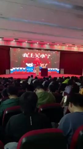 The scene shows a person onstage, dressed in a white robe and tall hat, at an event in a large auditorium, with a screen behind them displaying cartoon bears, and text that says, "I'm going to college." The person speaks to an audience seated in rows of red chairs. Many audience members are on their phones. The auditorium has bright lighting and red curtains at the back. There is a small table on the stage to the right of the person speaking.