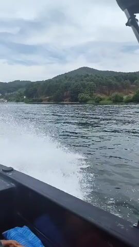 The video shows a speedboat cruising on a river. The camera is positioned inside the boat, facing forward and to the side. A person is visible in the foreground, wearing a black shirt and sunglasses. In the background, lush green hills covered in trees line the riverbanks. The sky is cloudy. The boat is creating a white wake as it moves through the water. Another person, a child wearing a blue hat and a dark swimsuit, is seated in front of the person in the foreground. They are both looking out at the scenery. The video appears to be taken from a personal perspective during a boat trip.