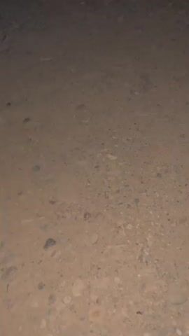 The video shows a grainy, low-light view of a street. In the background, there are several parked cars, a delivery truck, and a multi-story building with illuminated windows. The sky is dark, suggesting it is nighttime. The foreground is primarily composed of dirt and gravel.