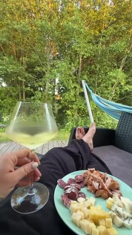 A person's hand holds a wine glass filled with white wine. In the foreground, a plate holds sliced meats and cheeses. The person is wearing black pants. Their bare feet are propped up, resting on an outdoor chair. Behind them, a hammock is visible. The scene is set outdoors, with lush green trees and foliage in the background, suggesting a natural, wooded environment. The lighting indicates it is likely late afternoon or early evening.