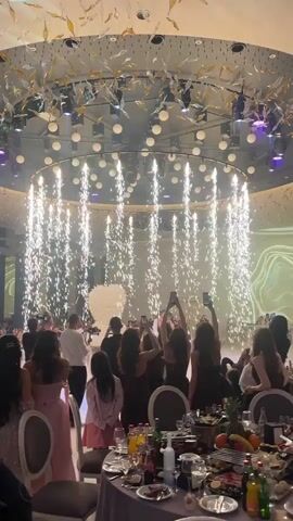 People are attending a wedding celebration. A bride and groom are on a stage in front of a white floral decoration, kissing. There are many guests surrounding them, most holding up cell phones to record the moment. Behind them is a circle of lights suspended from the ceiling that is shooting down sparks. On the lower right, there is a table with fruit and glasses.