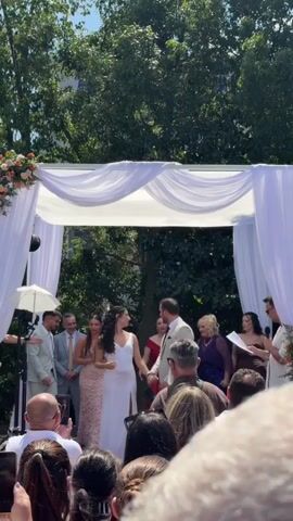 A wedding ceremony is underway outdoors under a white canopy adorned with draped fabric and floral arrangements. In the center, a bride in a white dress and a groom in a tan suit hold hands. They are surrounded by wedding party members and guests. The groom appears to be speaking into a microphone. The environment is sunny and appears to be a park or garden with trees in the background. Many guests are holding up phones, presumably to record the event. The overall atmosphere is celebratory.