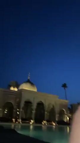 Here is a description of the video:

The video starts with a still shot of a building with a dome at night, illuminated by lights. In the foreground is a pool, also illuminated by lights, creating reflections on the water. The sky is a dark blue. The camera then moves to show a person's legs. The person is wearing a white lace dress. The camera continues to move, showing the area around the person, which appears to be a covered seating area with pillars and curtains. The lighting is dim, with a few candles providing some light.
