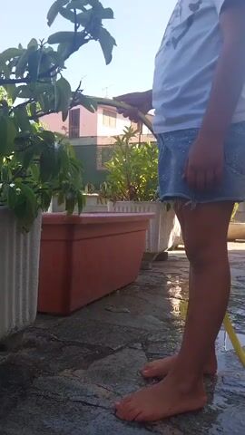 A person's bare feet and legs are visible from the knees down. The person is wearing a white t-shirt and a denim skirt. They are holding a yellow hose with a blue stripe and spraying water on a plant. The person is standing on a wet, stone patio. There are several planters around the person, one of which is a large, rectangular, reddish-brown planter. In the background, there are more planters and some buildings. The sun is shining, and the sky is blue.