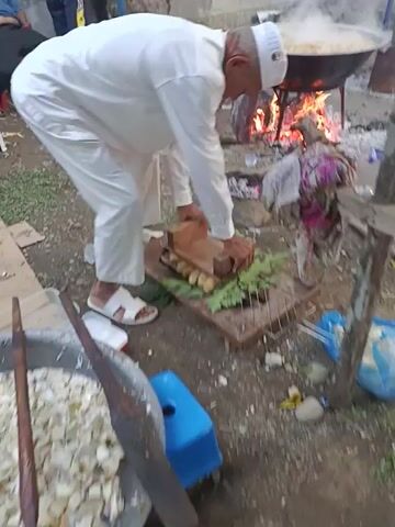 An overhead shot shows a man preparing a dish in an outdoor setting. The man, dressed in white and wearing a white skullcap, is kneeling. In front of him is a grill with several cooked bananas and green leaves. He is placing a wooden structure around the food on the grill. To the left, a fire burns inside a metal container. A woman is sitting in the background. To the right is a large pan containing a dish. The ground is covered with rocks and dirt. There are various containers and objects scattered around the area.