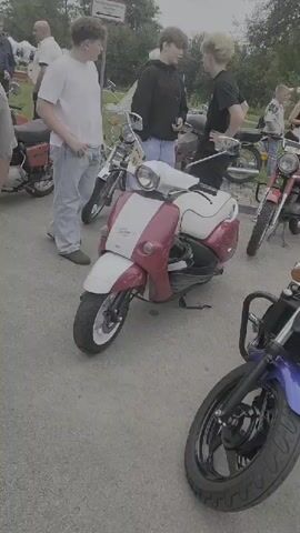 The video shows an outdoor gathering of people and motorcycles. In the foreground, a white and red scooter is parked. To the right of the scooter, a blue motorcycle is prominently displayed, with a man and a boy examining it. Further back, various other motorcycles are visible, including a red one to the left of the blue motorcycle and a maroon one to the right. People are scattered throughout the scene, some standing and talking, others appearing to interact with the motorcycles. The environment appears to be a paved area, possibly a parking lot or gathering space, with some greenery and a building in the background. The overall impression is of a casual event, likely a motorcycle show or rally.