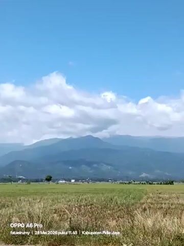 The video shows a landscape with mountains in the background and a field in the foreground. The sky is blue with scattered white clouds. In the distance, there are a few buildings, including what appears to be a house.  A few trees dot the landscape. The field in the foreground consists mostly of tall, dry grass.  The footage is steady. At the bottom of the video are text overlays indicating the camera model (OPPO A6 Pro), the name fataelrumy, and the date and time: 28 Mei 2026 pukul 11.43, as well as the location, Kabupaten Pidie Jaya.
