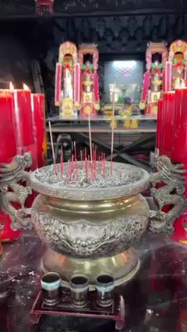 The video shows a close-up of a traditional Chinese shrine. In the foreground, there is a large, ornate metal incense burner filled with ashes and several lit incense sticks. In front of the burner, there are three small cups. To the left of the burner, there are large, lit red candles with golden Chinese characters on them. In the background, there are three elaborately decorated figures on a shelf, possibly deities or ancestors. The overall lighting is dim, and the focus is on the religious items.