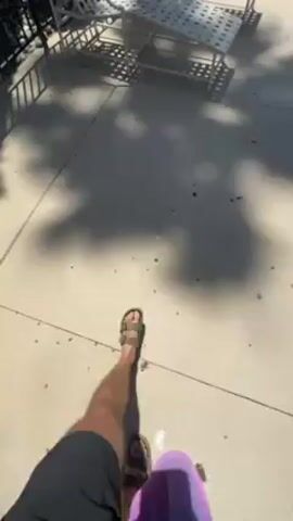 The video opens with a first-person view of a person's feet, wearing sandals, on a light-colored paved surface. A purple item is held in one hand, and the ground is dappled with shadows from trees and perforated outdoor furniture.

The camera then pans up to reveal a sunny outdoor swimming pool area. A rectangular pool with clear turquoise water and steps is the central feature. It is surrounded by beige paving. Numerous tall palm trees are prominent, their fronds swaying under a bright blue sky with scattered wispy clouds. The sun shines intensely, creating sharp shadows and occasionally appearing as a bright glare through the palm fronds.

The pool area includes white metal lounge chairs with a perforated pattern, and at one end, a white umbrella shades a table. The perimeter of the area features a black metal fence, behind which are well-maintained green shrubs and bushes with purple flowers. In the background, houses with reddish-brown roofs are visible, nestled among more palm trees and greenery. The scene conveys a peaceful and warm atmosphere, characteristic of a sunny day in a resort or residential community.