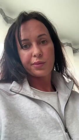 A woman with long, dark hair is holding a wine glass filled with a light-colored liquid. She is wearing a grey zip-up sweatshirt over a white t-shirt. The background appears to be indoors, with some soft lighting and what looks like curtains or blinds. The woman looks directly at the camera with a slight smile.
