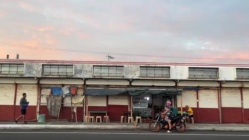 The video captures a street scene with a building in the background. The building appears to be a row of shops with roll-up doors, many of which are closed. The upper part of the building is painted white with horizontal windows. The lower part is painted red. There is a small open stall in front of one of the shops with a tarp for cover, and several stools are set up. A person on a motorcycle with a passenger is stopped in front of the stall. To the left, a person is standing near some laundry hanging on a makeshift rack. Further to the left, a person is standing near a green bucket. To the right, a person is sitting on a stool. The sky above is partly cloudy with pink and orange hues, suggesting it might be sunrise or sunset. The street in the foreground is paved. Power lines are visible against the sky.