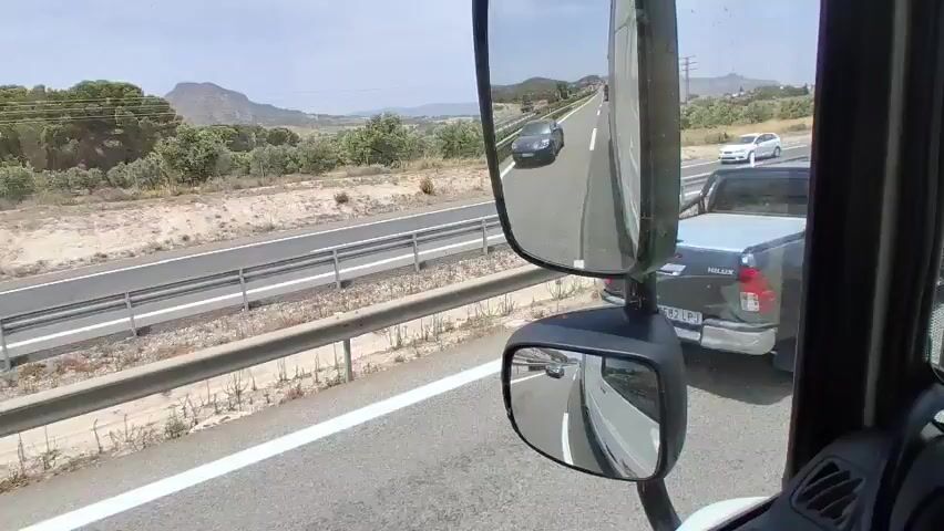 The video shows a view from inside a vehicle, likely a truck, looking forward and to the side. The vehicle is on a highway with multiple lanes. In the foreground, a large side mirror reflects the road and other vehicles. Outside the vehicle, a black sports car is visible in the lane to the left. Further down the highway, other vehicles including a white van and a pickup truck are also present. The landscape on either side of the highway is dry and hilly with sparse vegetation. The sky is clear and the lighting suggests it is daytime. The video captures the movement of traffic on the highway from the perspective of the driver or passenger of the forward-moving vehicle.